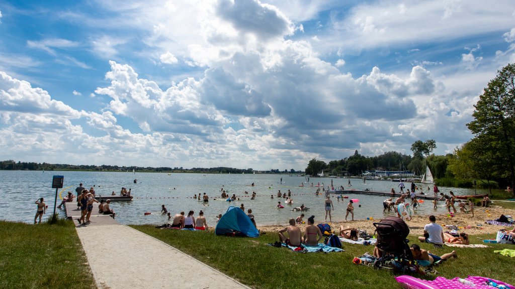 Zdjęcie przedstawia ludzi siedzących na plaży i kąpiących się w jeziorze.