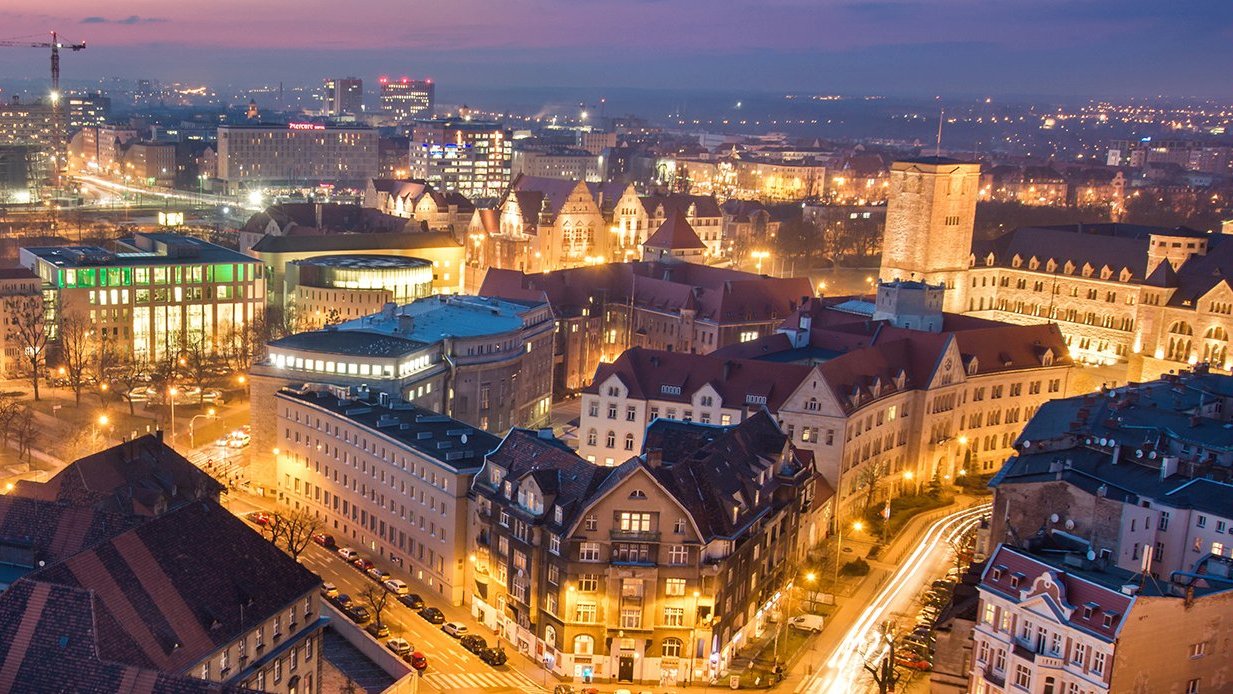 Grafika: panorama Poznania po zachodzie słońca, na pierwszym planie kamienice, w tle CK Zamek