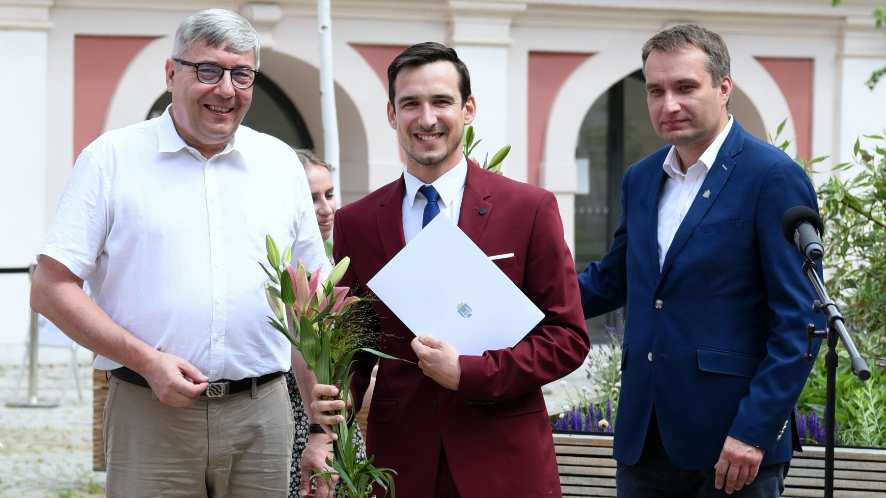 Dziedziniec Urzędu Miasta Poznania. Stypendysta trzymający w ręku kwiat i teczkę, obok niego Grzegorz Ganowicz i Mariusz Wiśniewski