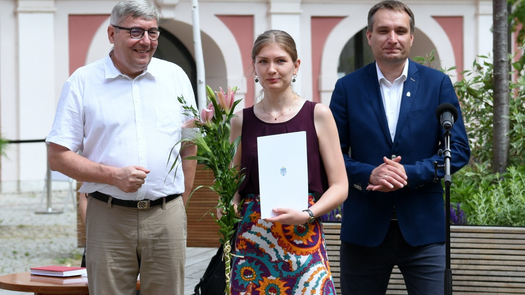 Dziedziniec Urzędu Miasta Poznania. Stypendystka trzymający w ręku kwiat i teczkę, obok Grzegorz Ganowicz i Mariusz Wiśniewski