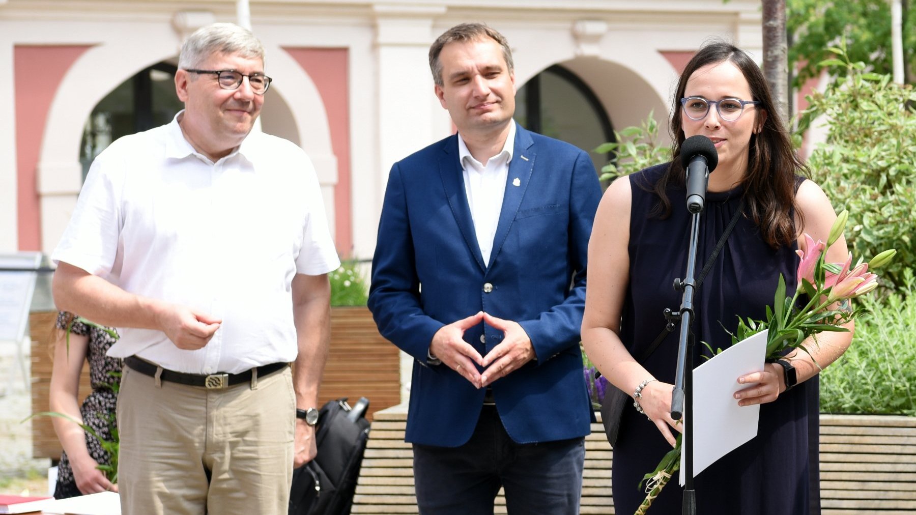 Dziedziniec Urzędu Miasta Poznania. Stypendystka trzymająca w ręku kwiat i teczkę, przy mikrofonie, obok niej Grzegorz Ganowicz i Mariusz Wiśniewski