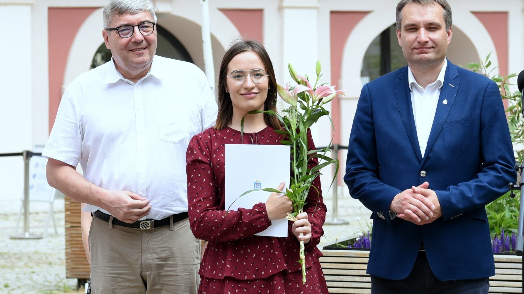 Dziedziniec Urzędu Miasta Poznania. Stypendystka trzymająca w ręku kwiat i teczkę, obok niej Grzegorz Ganowicz i Mariusz Wiśniewski