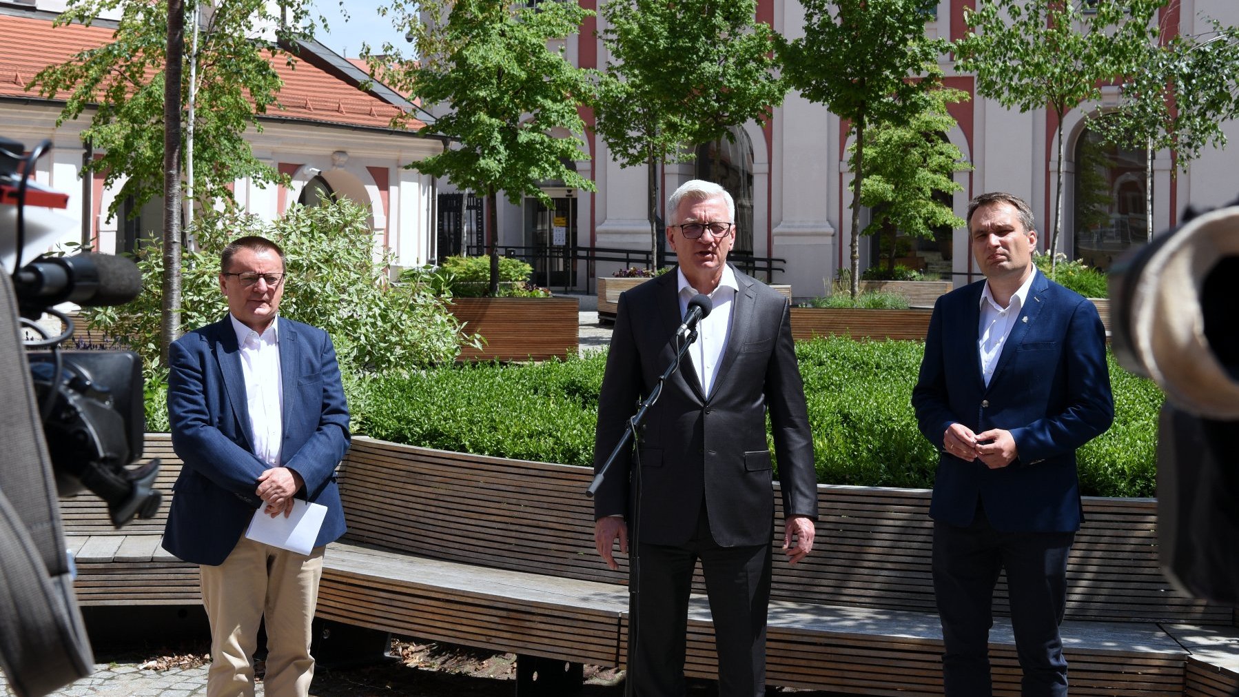 Na zdjęciu dziedziniec urzędu miasta, przy mikrofonach stoi trzech mężczyzn, w centrum Jacek Jaśkowiak, obok Mariusz Wiśniewski, w tle drzewa