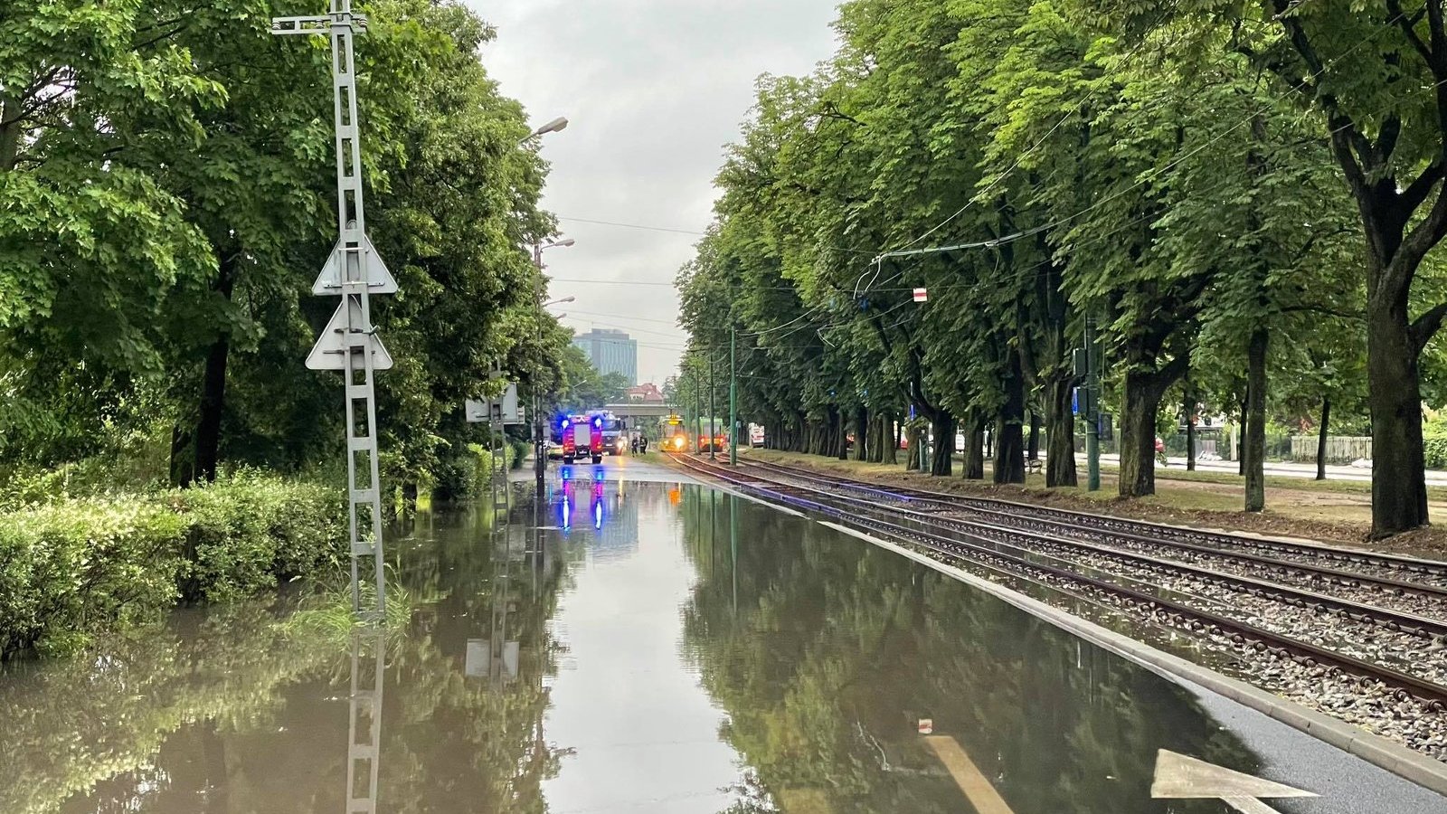 Na zdjęciu zalana ulica, obok torowisko. Po bokach drzewa, w tle samochód straży pożarnej na sygnałach