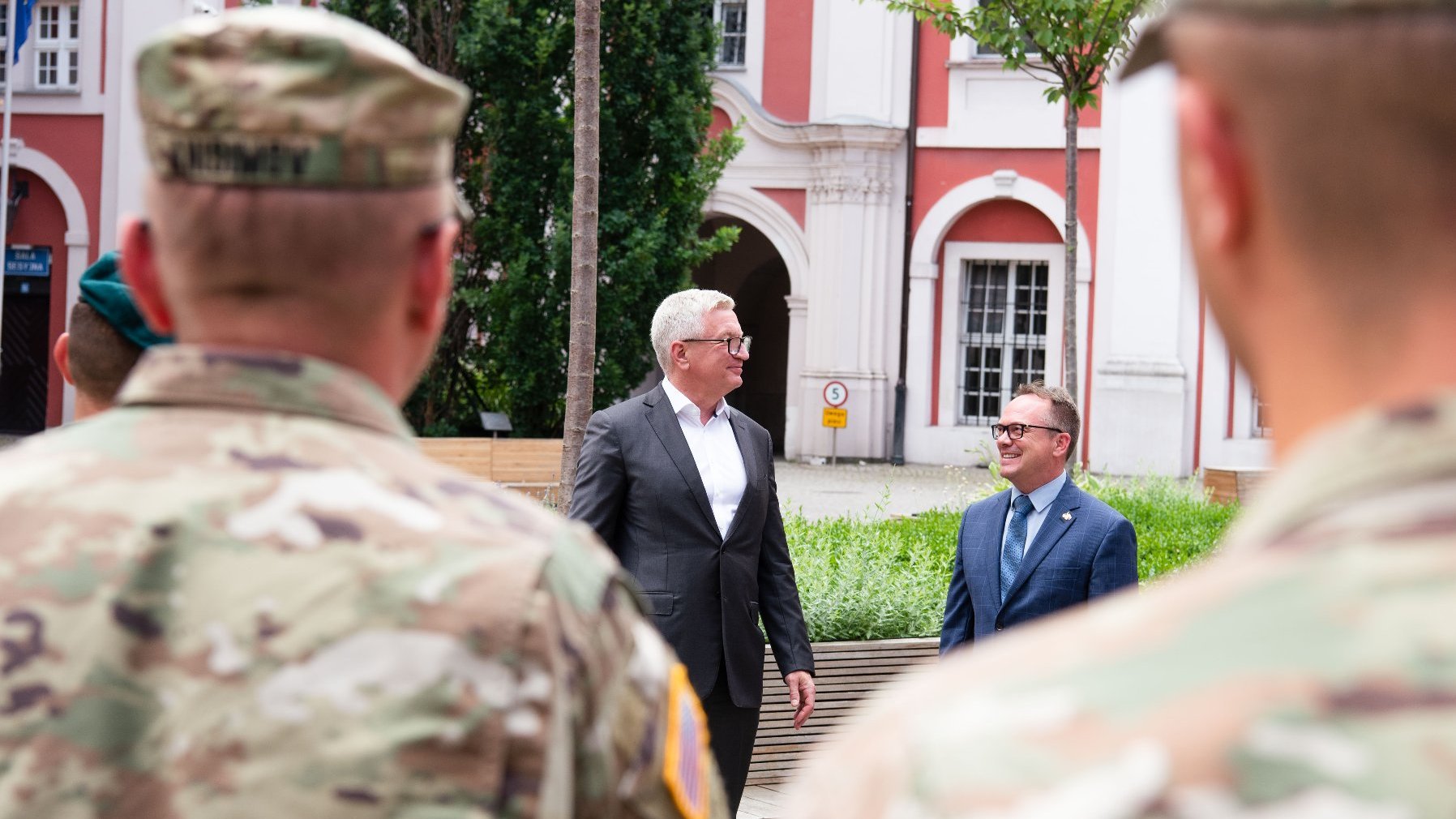 Dziedziniec Urzędu Miasta Poznania. Jacek Jaśkowiak i Scott Brokaw patrzą na siebie, uśmiechnięci. Na pierwszym planie, nieostrym - żołnierze stojący tyłem do obiektywu