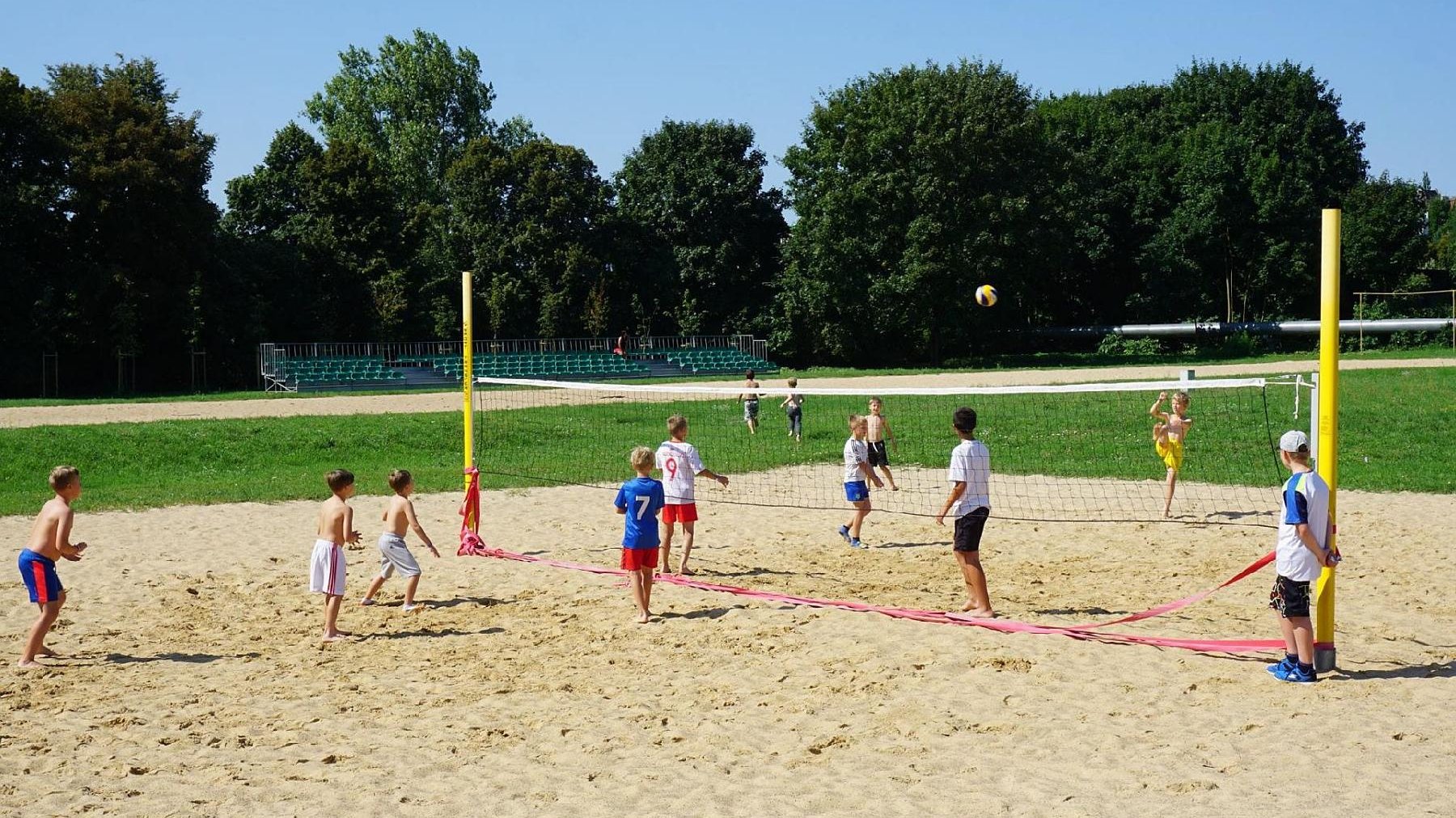 Zdjęcie: boisko do siatkówki plażowej na którym gra kilkoro dzieci, w tle szpaler drzew - grafika rozmowy