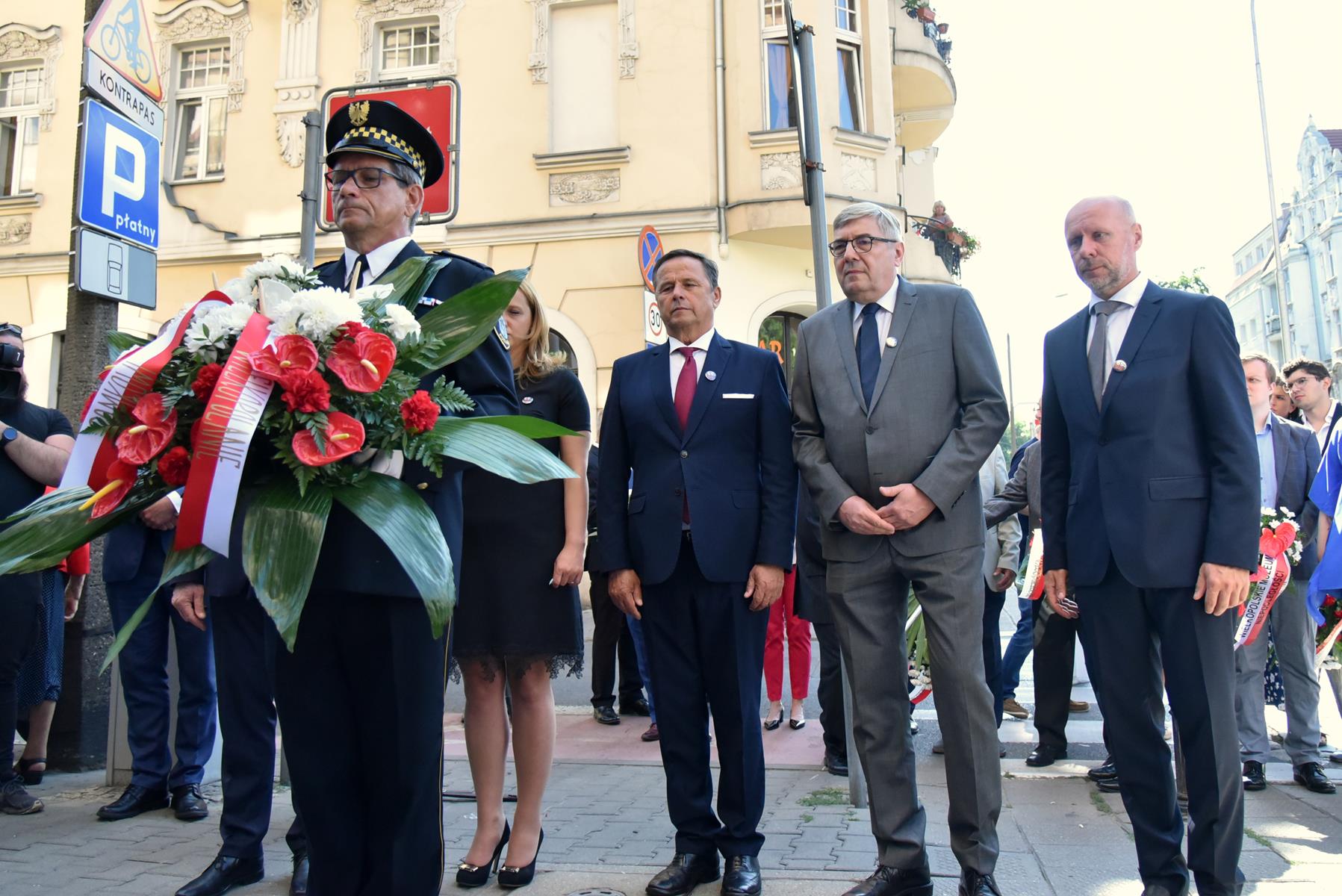 Na zdjęciu grupa ludzi składających wiązankę, wśród nich Grzegorz Ganowicz i Bartosz Guss, na pierwszym planie strażnik z kwiatami - grafika rozmowy