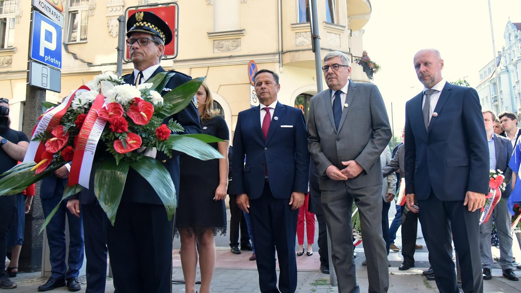 Na zdjęciu grupa ludzi składających wiązankę, wśród nich Grzegorz Ganowicz i Bartosz Guss, na pierwszym planie strażnik z kwiatami