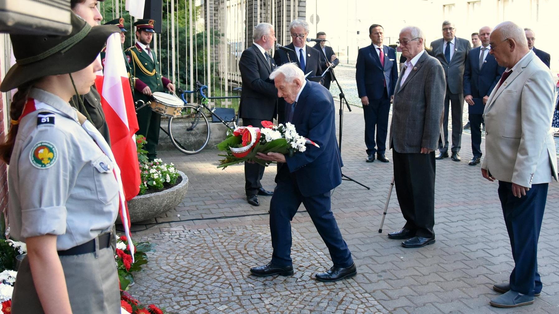 Składanie kwiatów. Po lewej harcerze, po prawej grupa mężczyzn, w środku starszy człowiek z wiązanką - Zenon Wechmann