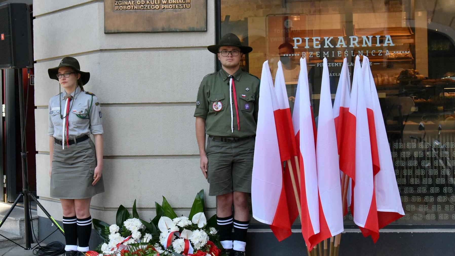 Tablica pamięci Romka Strzałkowskiego, obok harcerze i polskie flagi, pod tablicą kwiaty