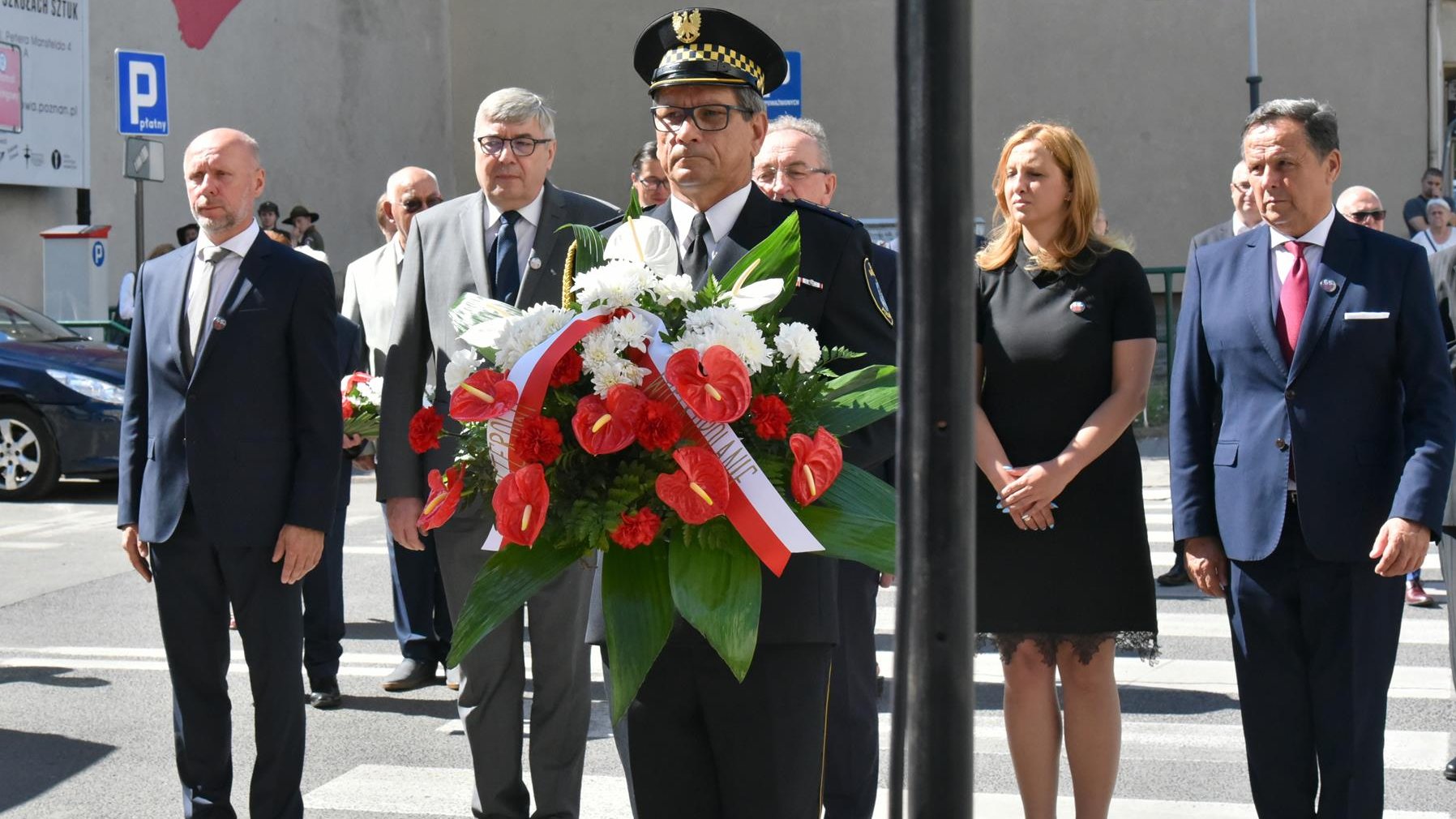 Grupa ludzi składających kwiaty, stojących w rzędzie, wśród nich Bartosz Guss i Grzegorz Ganowicz, na pierwszym planie strażnik z wiązanką