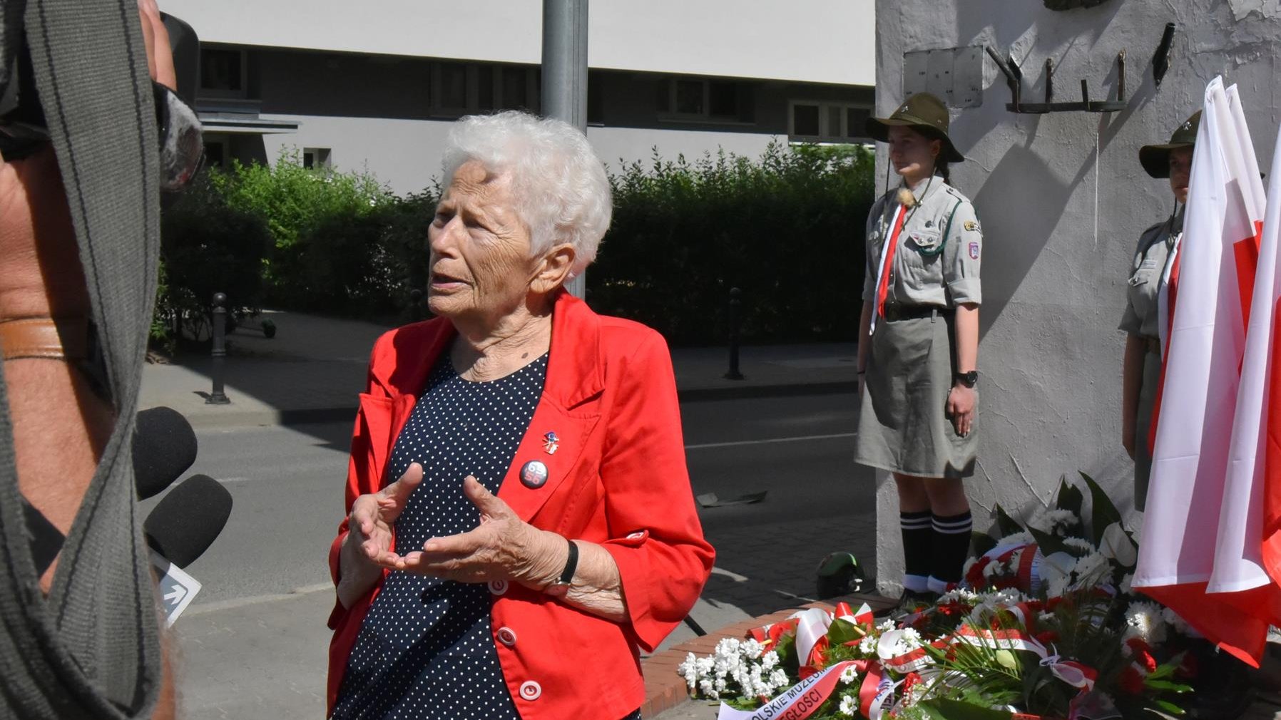 Kobieta w czerwonym żakiecie - Aleksandra Banasiak - gestukuluje, w tle tablica im. Petera Mansfelda