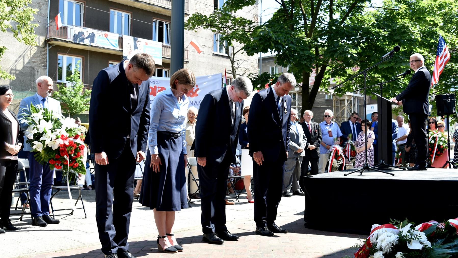 Grupa czterech osób skłania głowy, za nimi budynek mieszkalny, w tle banery Solidarności i flaga Polski