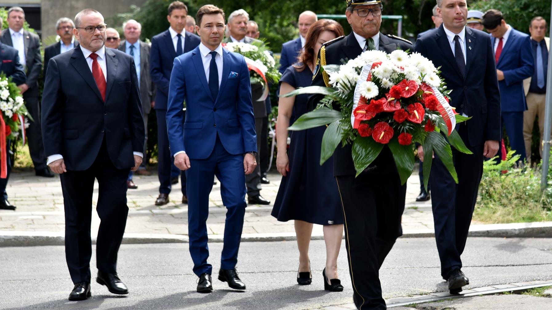 Grupa osób składa wiązankę, na pierwszym planie strażnik miejski z kwiatami