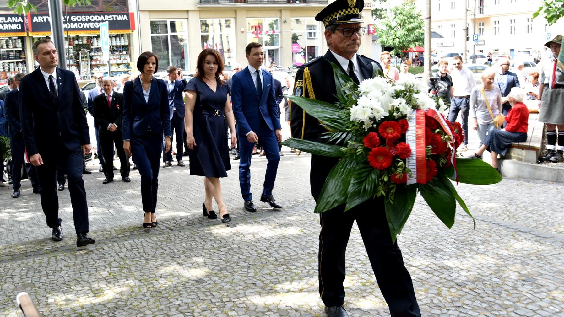 Cztery osoby idące obok siebie, dwie kobiety i dwóch mężczyzn, na pierwszym planie strażnik miejski z biało-czerwoną wiązanką