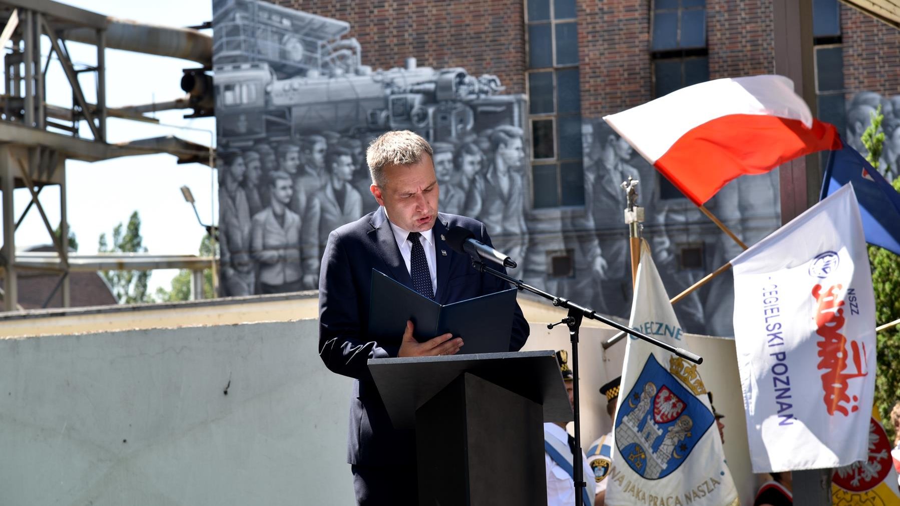 Mariusz Wiśniewski, zastępca prezydenta Poznania, przy mikrofonie, w tle mural, obok flagi Polski, Miasta Poznania i "Solidarności"