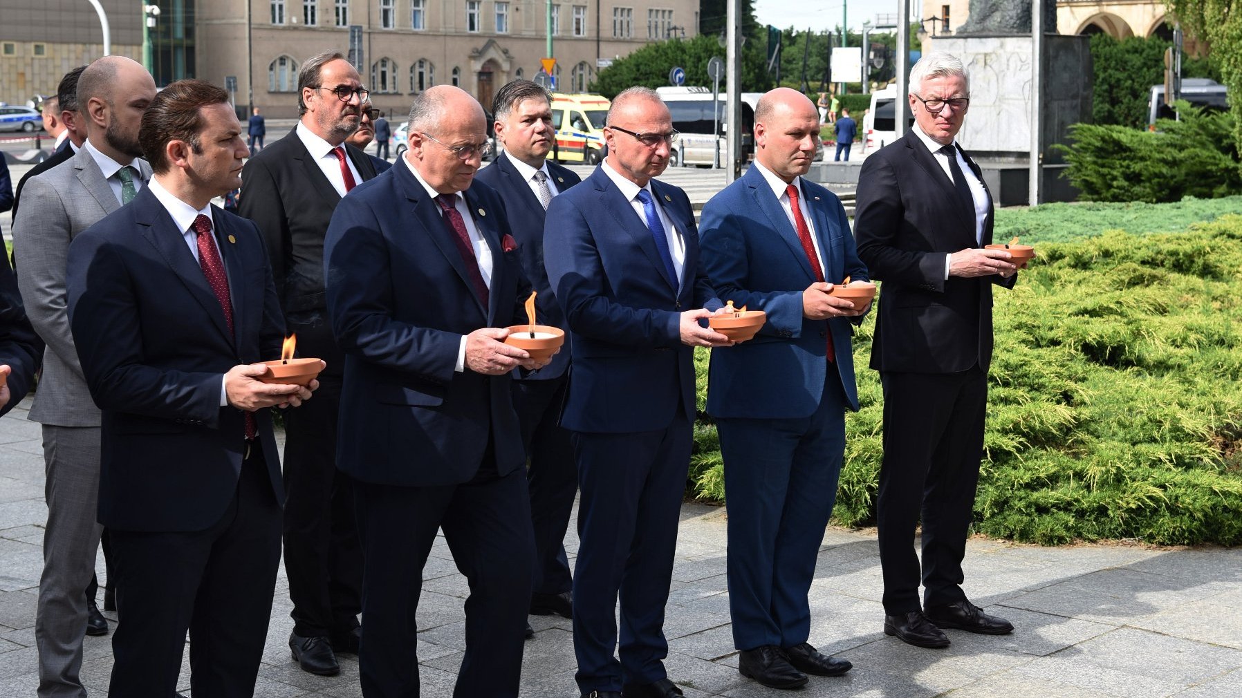 Grupa mężczyzn w garniturach, stoją w rzędzie, każdy trzyma duży znicz. Wśród nich Jacek Jaśkowiak, prezydent Poznania