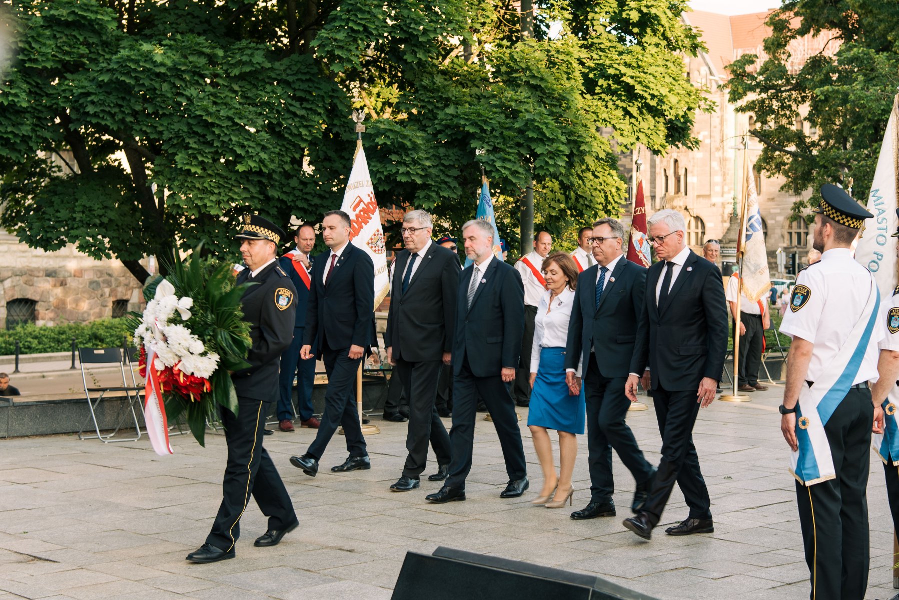 Na zdjęciu grupa samorządowców - wśród nich Jacek Jaśkowiak - składa kwiaty pod pomnikiem. Na pierwszym planie strażnik miejski z wiązanką - grafika rozmowy