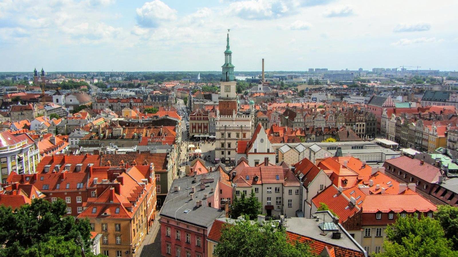 Panorama Poznania, na pierwszym planie dachy kamienic, w centrum wieża Ratusza, u góry błękitne niebo, na nim białe chmury - grafika rozmowy