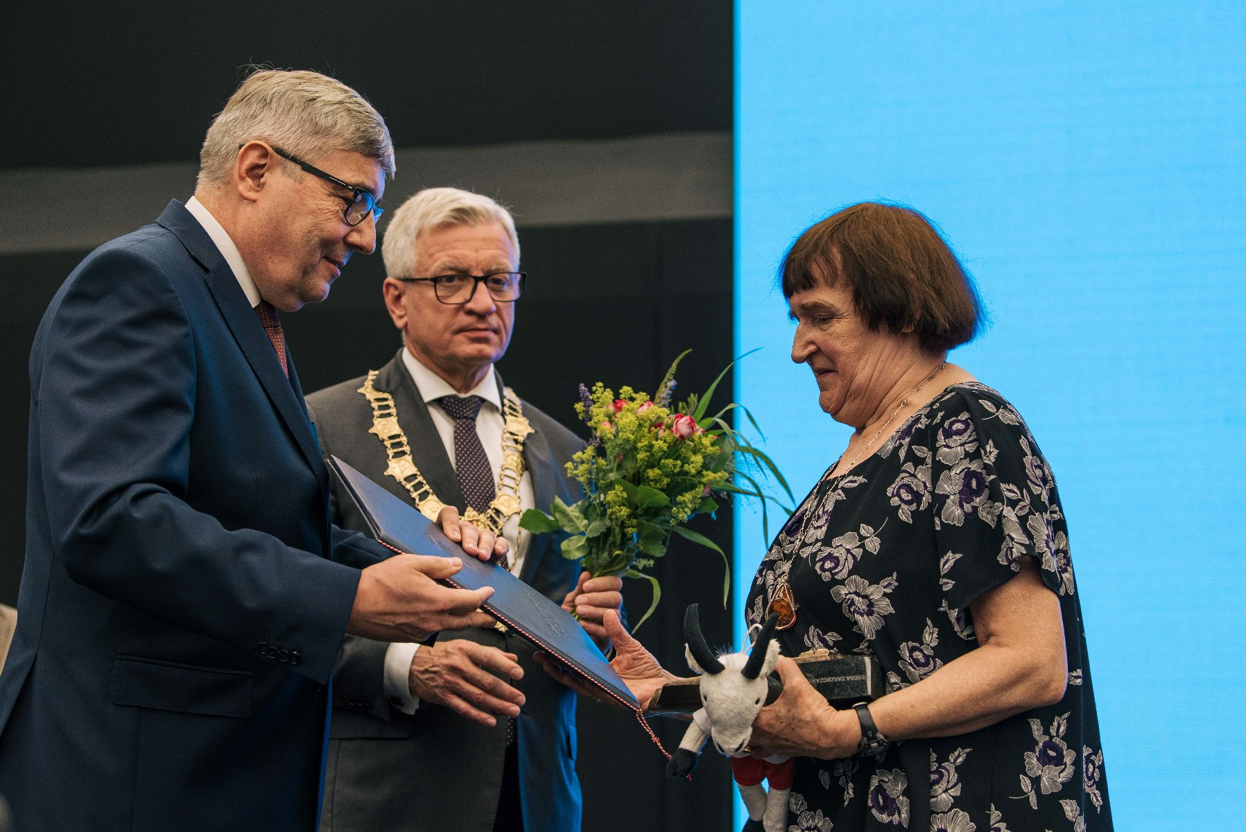 Kobieta odbierająca nagrodę z rąk Grzegorza Ganowicza, za nią Jacek Jaśkowiak trzymający kwiaty - grafika rozmowy