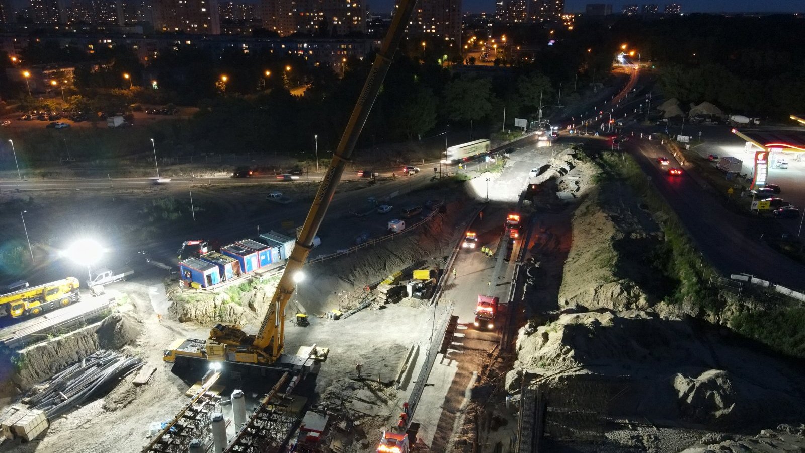 Na zdjęciu plac budowy nocą, w centrum duży dźwig i sznur samochodów