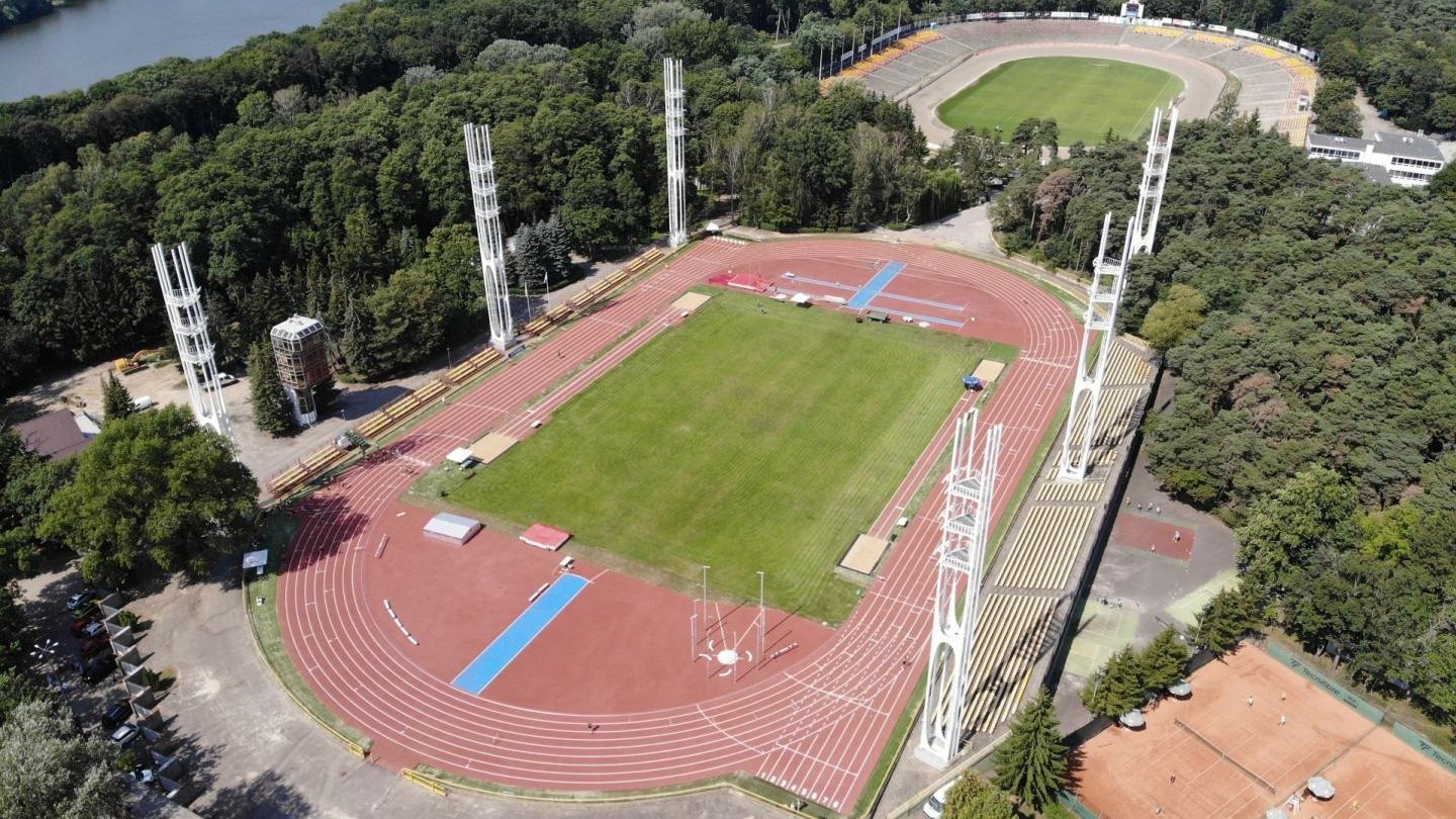 Widoka na stadion lekkoatletyczny z lotu ptaka. Wokół zieleń, w tle stadion piłkarski i jezioro Rusałka.