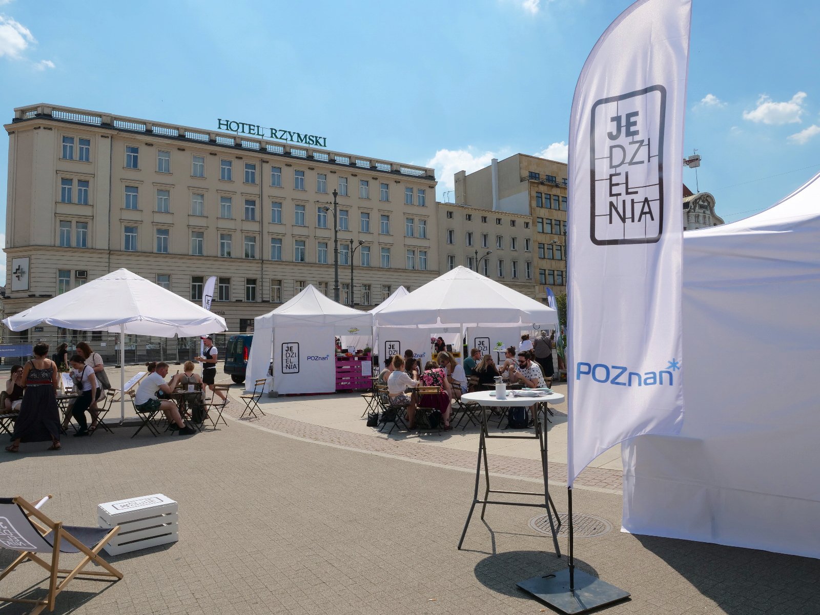 Zdjęcie przedstawia ludzi siedzących przy stolikach na Placu Wolności. Obok znajdują się białe namioty i parasole oraz baner z napisem "Jedzielnia". W tle widać Hotel Rzymski. - grafika rozmowy