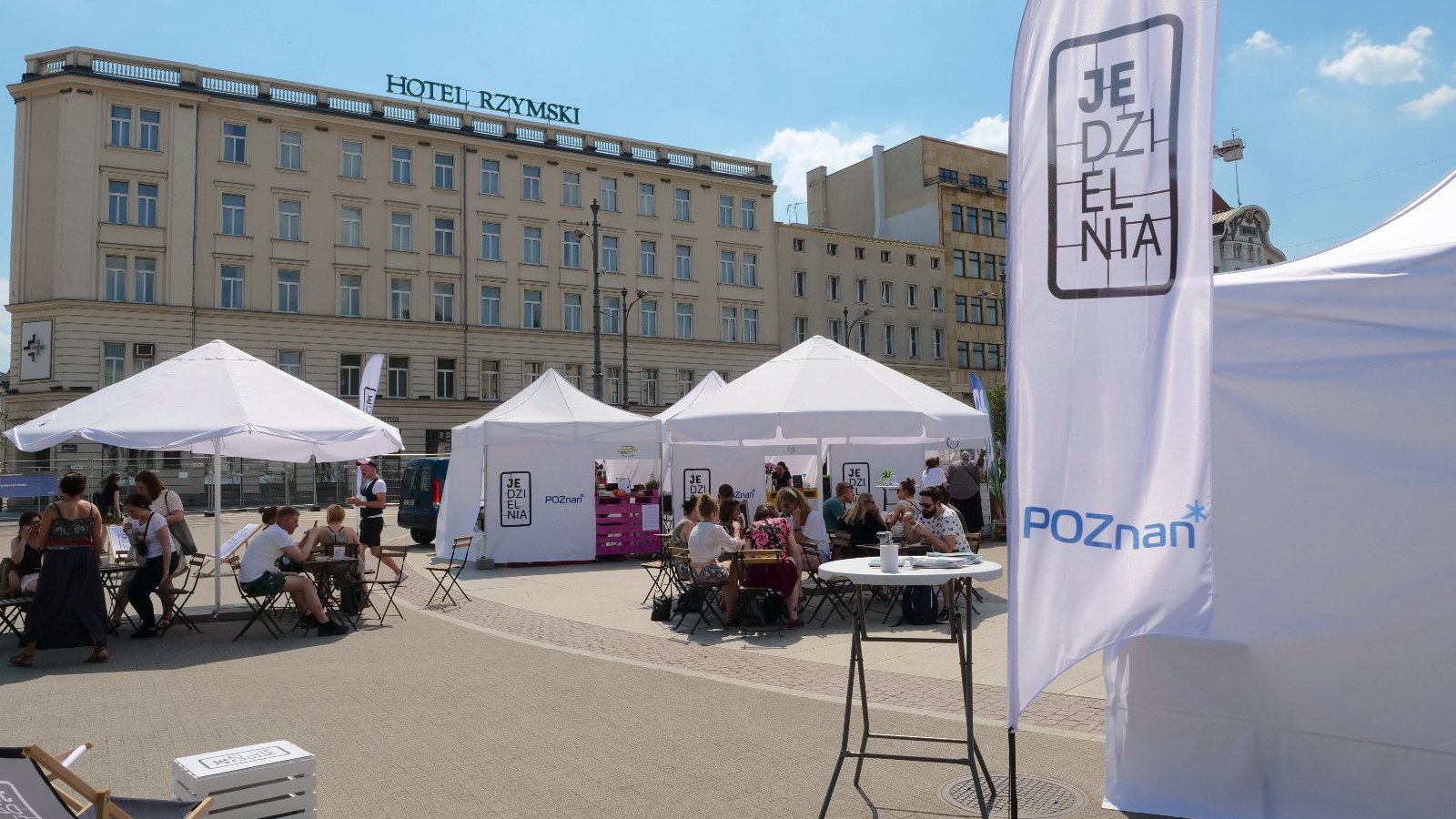 Zdjęcie przedstawia ludzi siedzących przy stolikach na Placu Wolności. Obok znajdują się białe namioty i parasole oraz baner z napisem "Jedzielnia". W tle widać Hotel Rzymski.