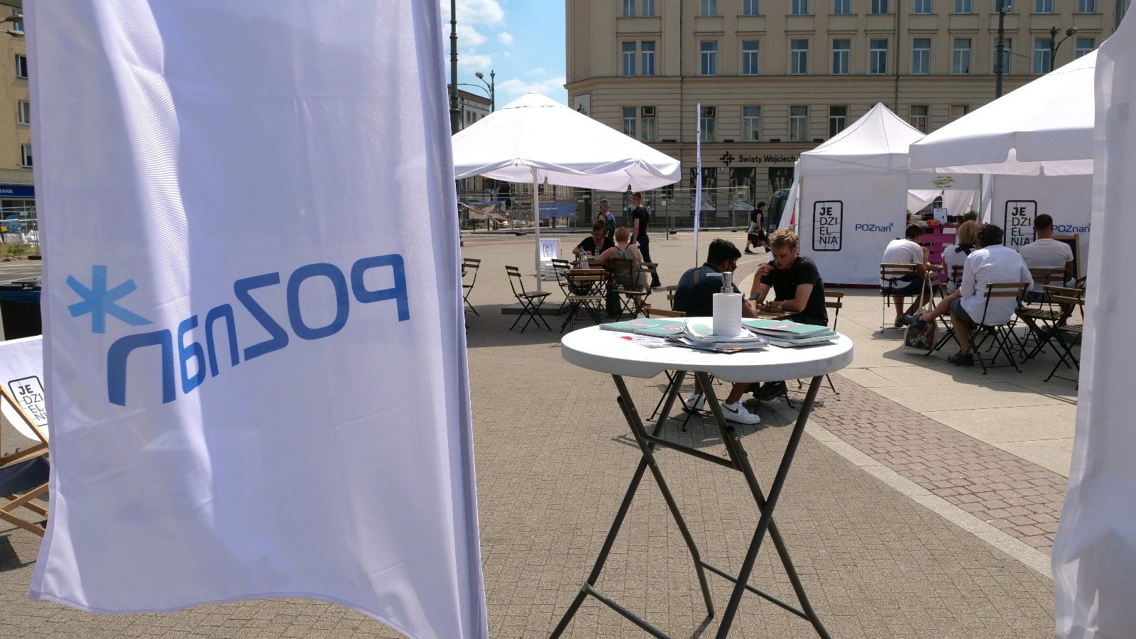 Zdjęcie przedstawia ludzi siedzących przy stolikach na Placu Wolności. W tle widać białe namioty z napisem "Jedzielnia" i biały parasol. Na pierwszym planie po lewej stronie widać baner z napisem "Poznań".