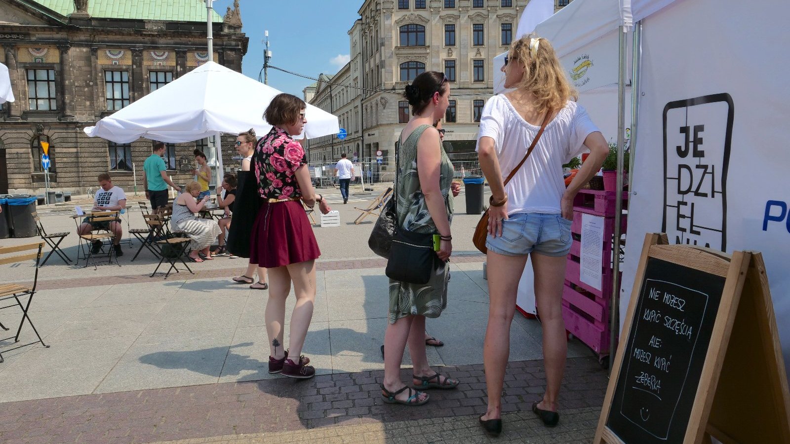 Zdjęcie przedstawia ludzi stojących w kolejce przed białym namiotem. W tle widać innych ludzi siedzących przy stoliku pod parasolem.