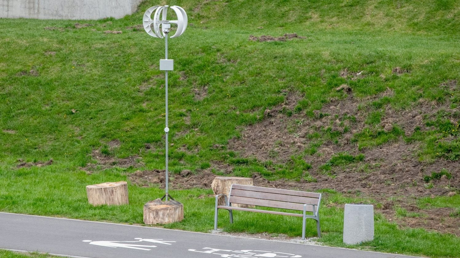 Zdjęcie przedstawia instalację artystyczną o nazwie Wiatrak. Obok znajduje się ławka i drewniane pieńki.