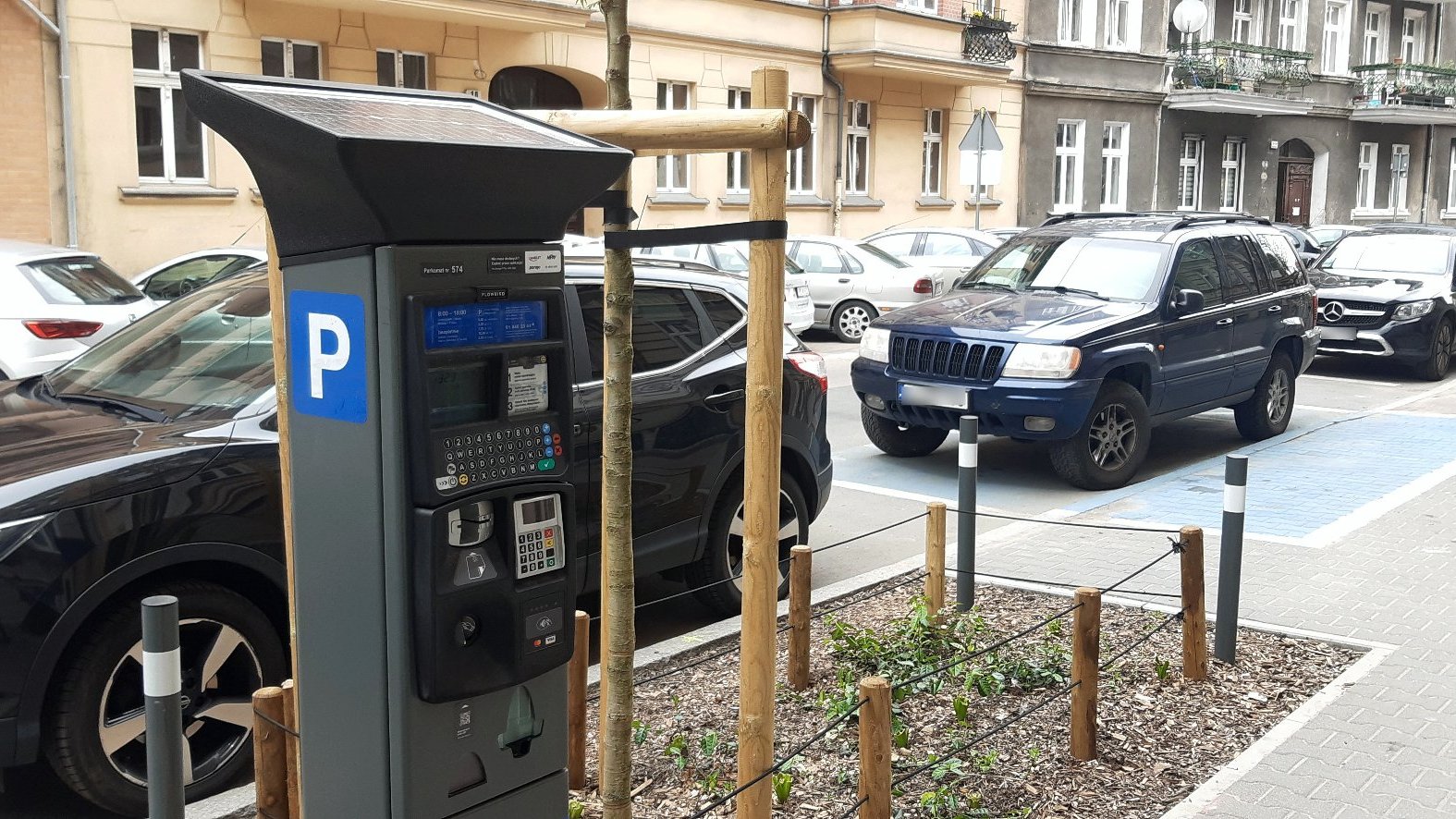 Zdjęcie ulicy, przy której stoją zaparkowane samochody. Na pierwszym planie parkomat, w tle kamienice - grafika rozmowy