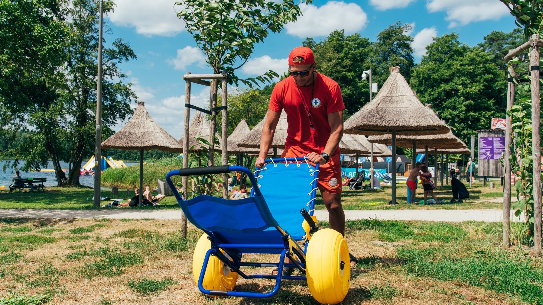 Zdjęcie przedstawia ratownika z wózkiem amfibią na plaży.