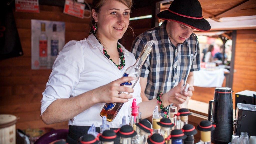 Zdjęcie przedstawia kobietę i mężczyznę w czarnym kapeluszu. Kobieta trzyma w ręku butelkę z nalewką.