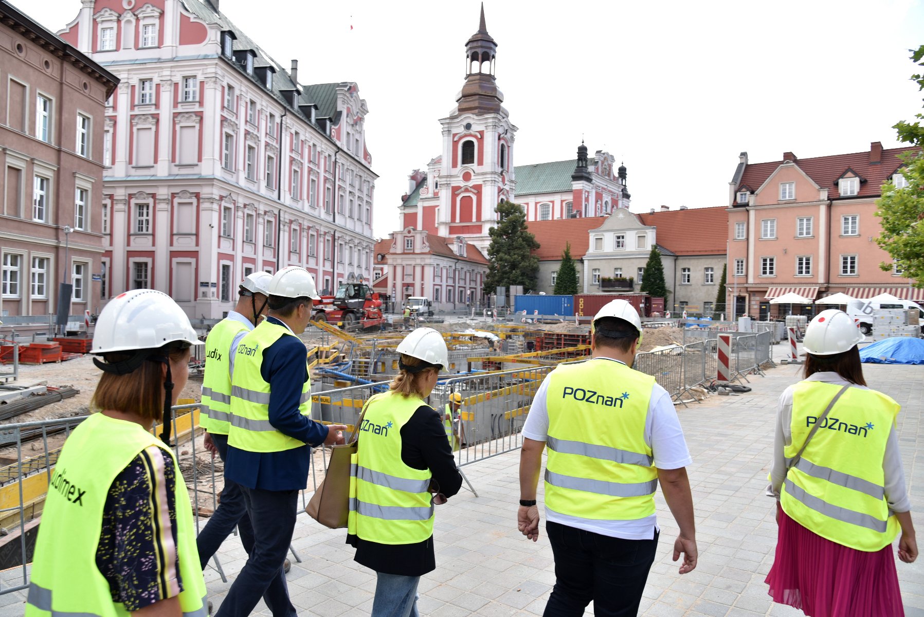 Plac Kolegiacki. Na pierwszym planie grupa osób w kaskach i kamizelkach odblaskowych. Na dalszym planie plac budowy. W tle budynek urzędu miasta. - grafika rozmowy