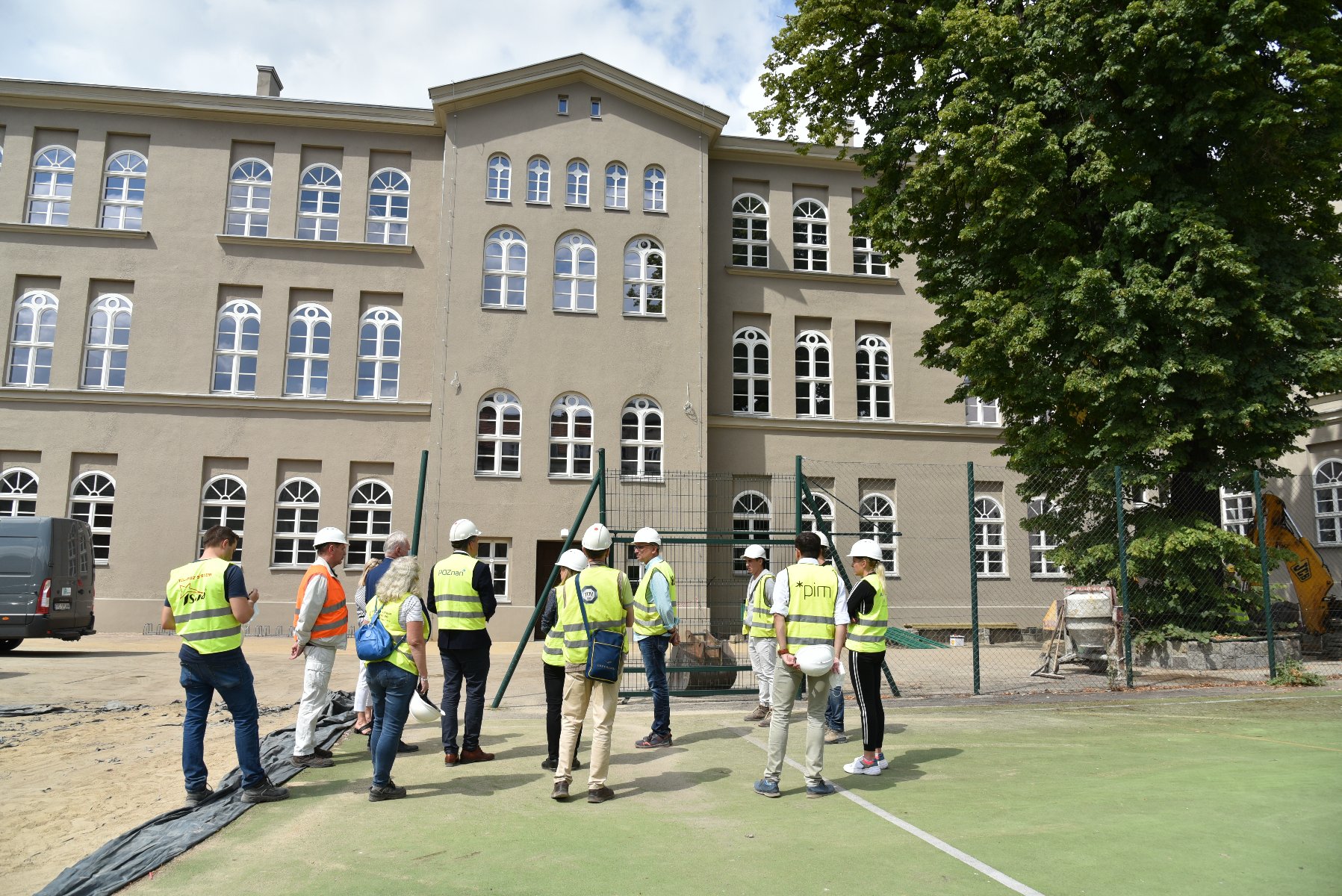 Zdjęcie przedstawia grupę ludzi w żółtych kamizelkach stojących przed remontowanym budynkiem szkoły. - grafika rozmowy