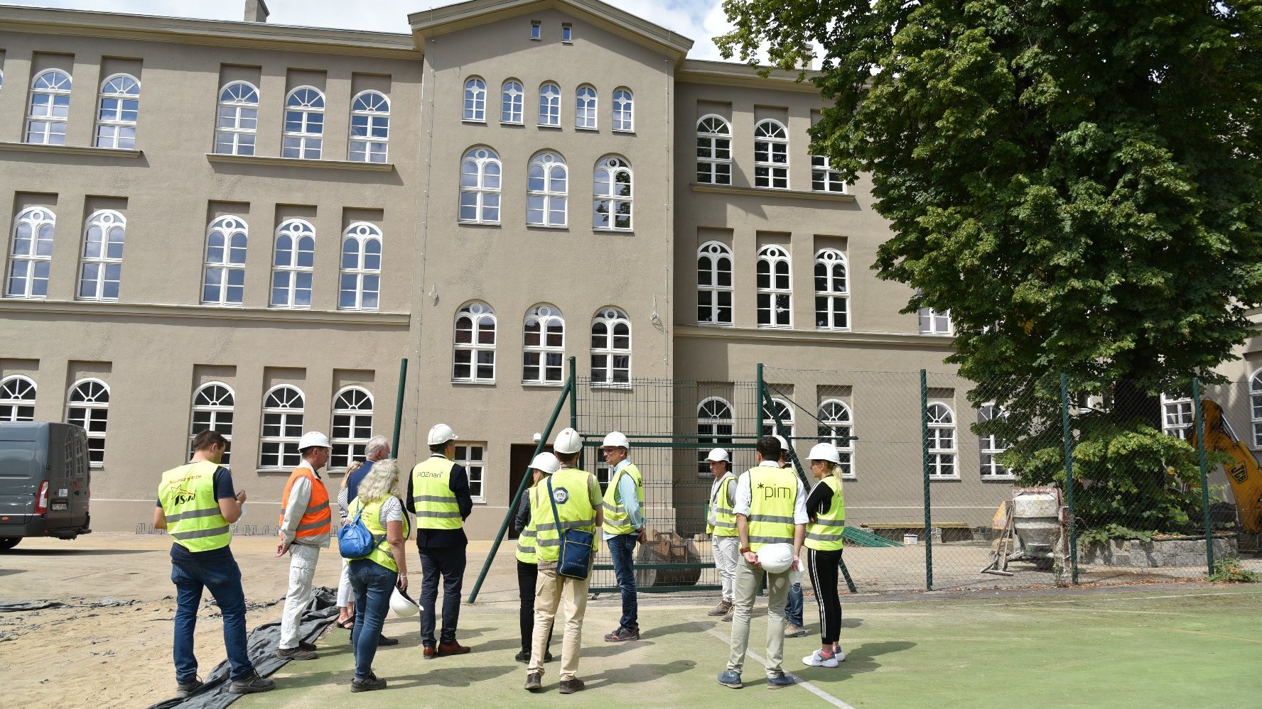 Zdjęcie przedstawia grupę ludzi w żółtych kamizelkach stojących przed remontowanym budynkiem szkoły.