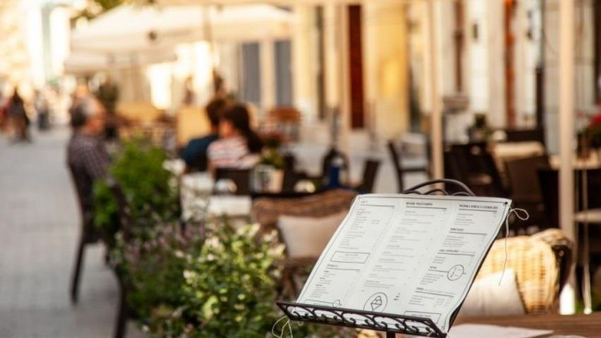 Na zdjęciu ogródek gastronomiczny, na pierwszym planie wystawione menu, w tle nieostre stoliki - grafika rozmowy