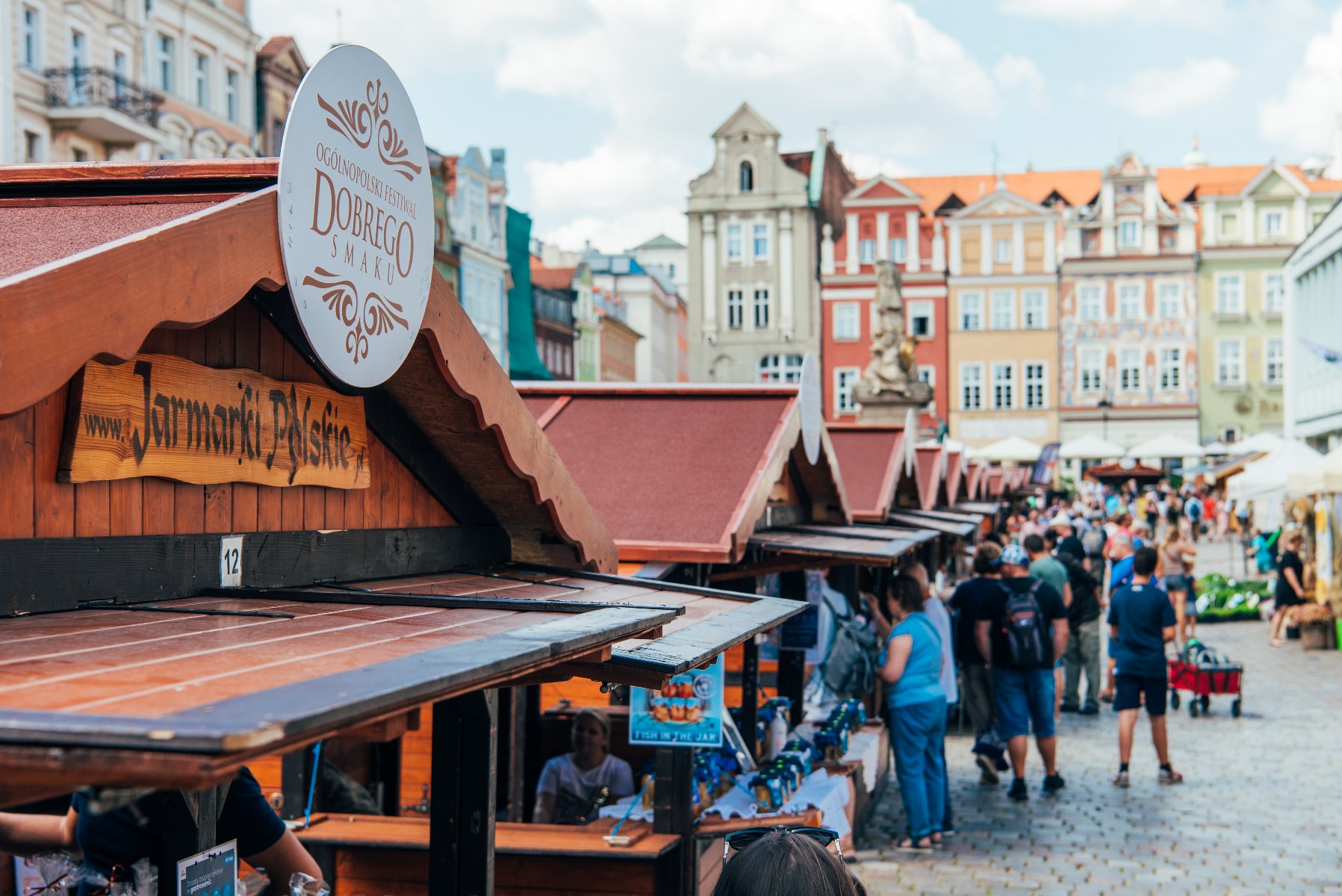 Zdjęcie przedstawia tłum ludzi na rynku. Gromadzą się wokół drewnianych domków. Na pierwszym planie widać logo z nazwą festiwalu. Jest przyczepione do dachu jednego z domków. - grafika rozmowy