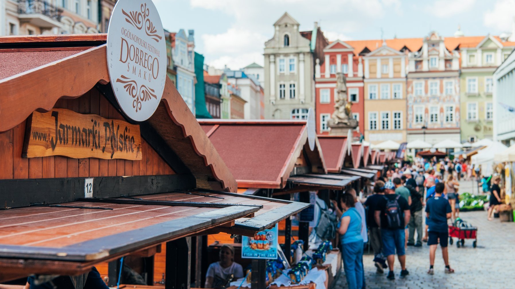 Zdjęcie przedstawia tłum ludzi na rynku. Gromadzą się wokół drewnianych domków. Na pierwszym planie widać logo z nazwą festiwalu. Jest przyczepione do dachu jednego z domków.