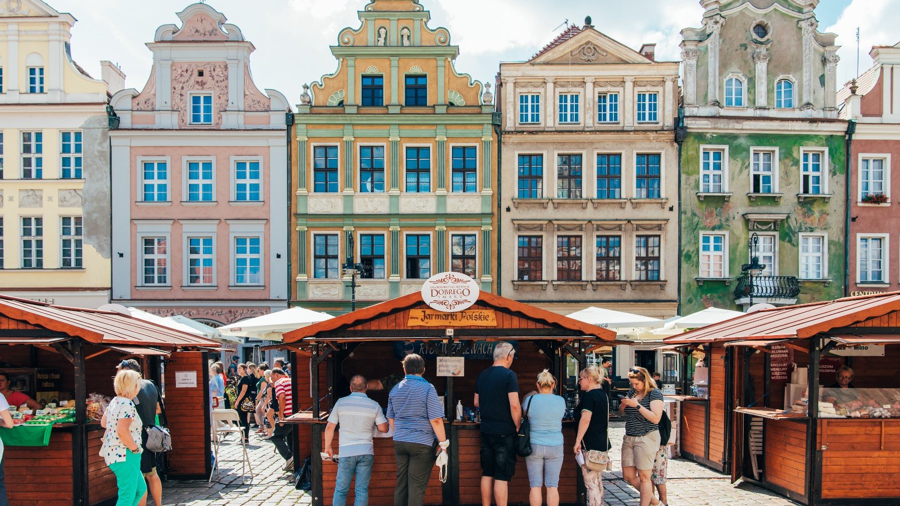 Zdjęcie przedstawia drewniane domki oraz stojących przy nich ludzi. W tle widać kolorowe kamienice.