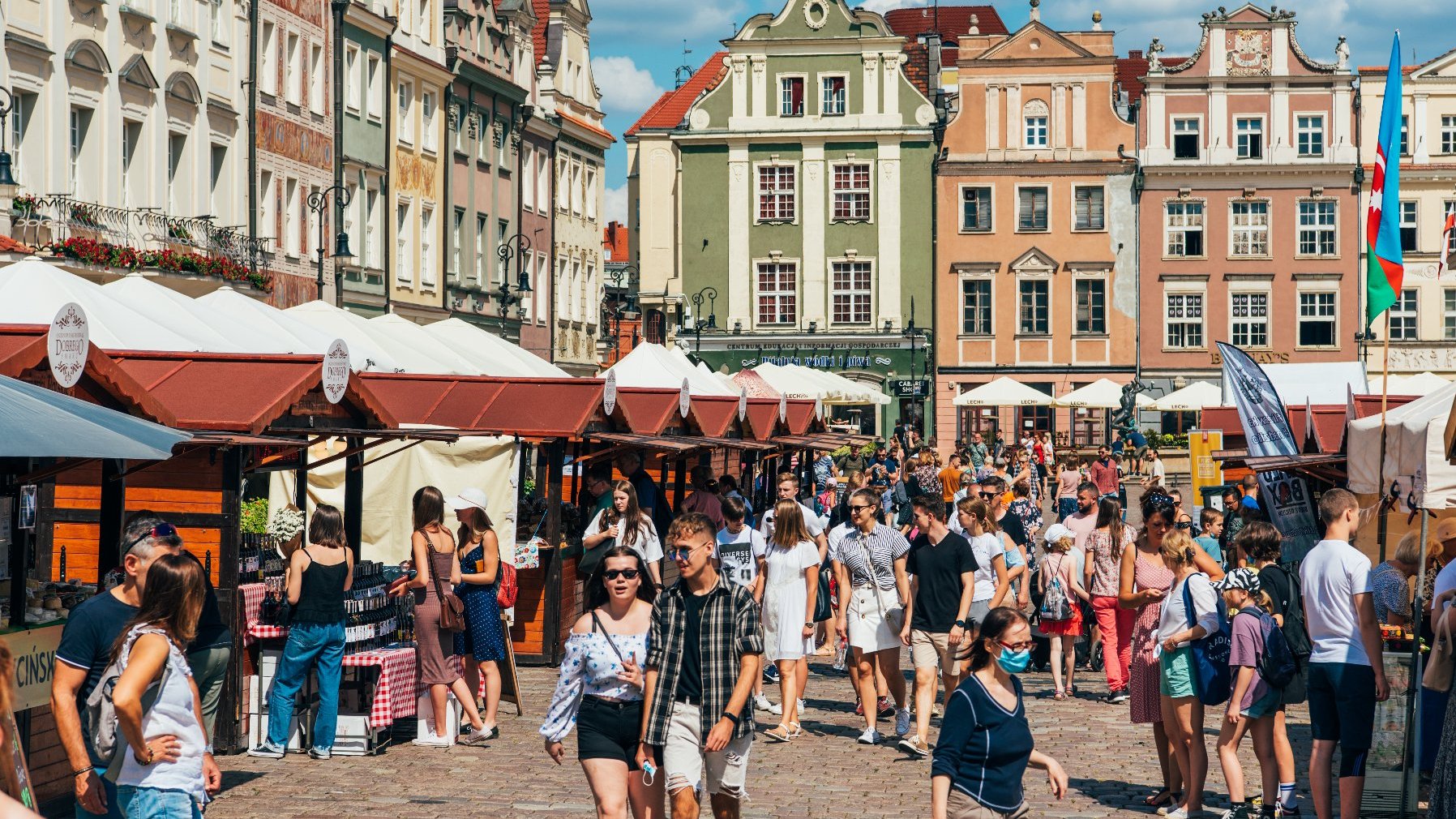 Zdjęcie przedstawia tłum ludzi na rynku. Obok widać stragany i kamienice.