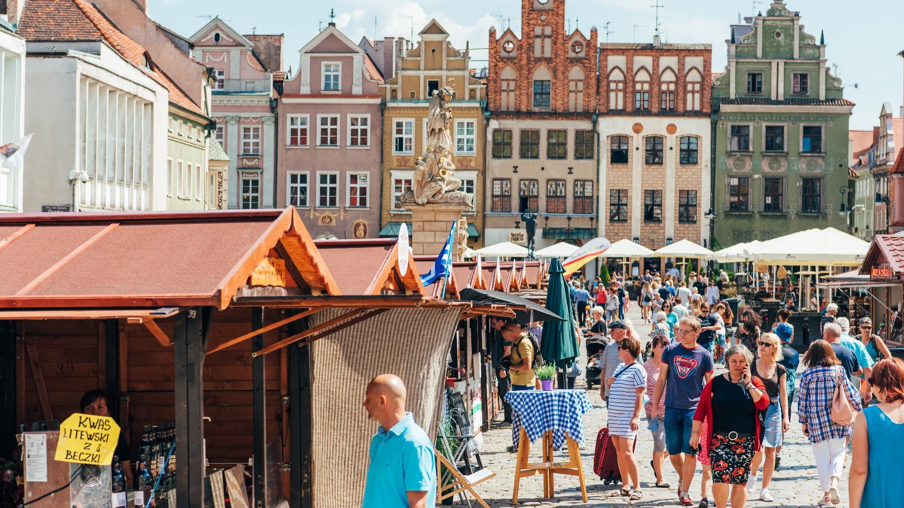 Zdjęcie przedstawia tłum ludzi na rynku, chodzących wokół drewnianych domków. W tle widać kamienice.