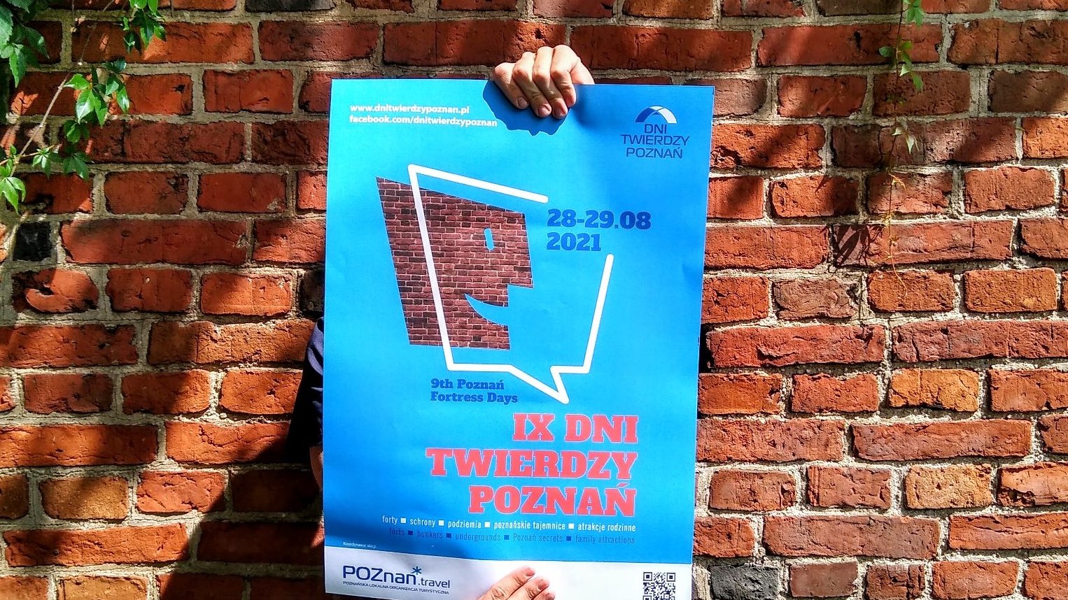 Zdjęcie przedstawia plakat IX Dni Twierdzy Poznań. Ktoś trzyma plakat, widać tylko jego dłonie. W tle znajduje się ściana z czerwonych cegieł.