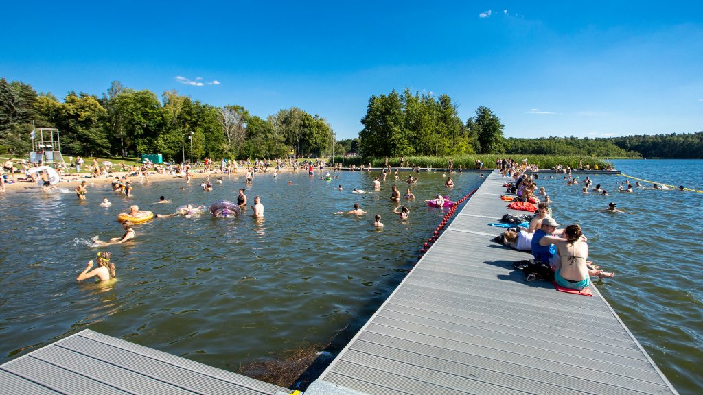 Zdjęcie przedstawia kąpielisko. Widać na nim pomost i wodę, w oddali także plażę. Wszędzie jest tłum ludzi.