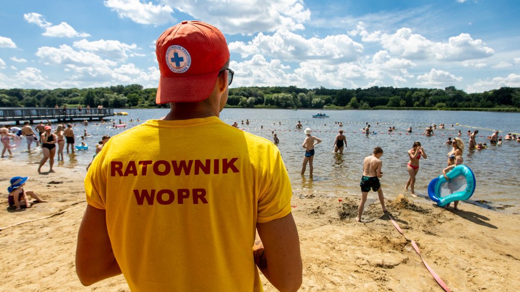 Zdjęcie przedstawia stojącego tyłem do aparatu ratownika. Dalej widać plażę i wodę, w której kąpią się ludzie.