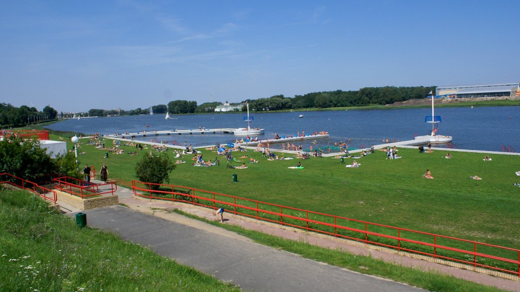 Zdjęcie przedstawia kąpielisko. Widać na nim trawiastą plażę, na której wypoczywają ludzie oraz jezioro.