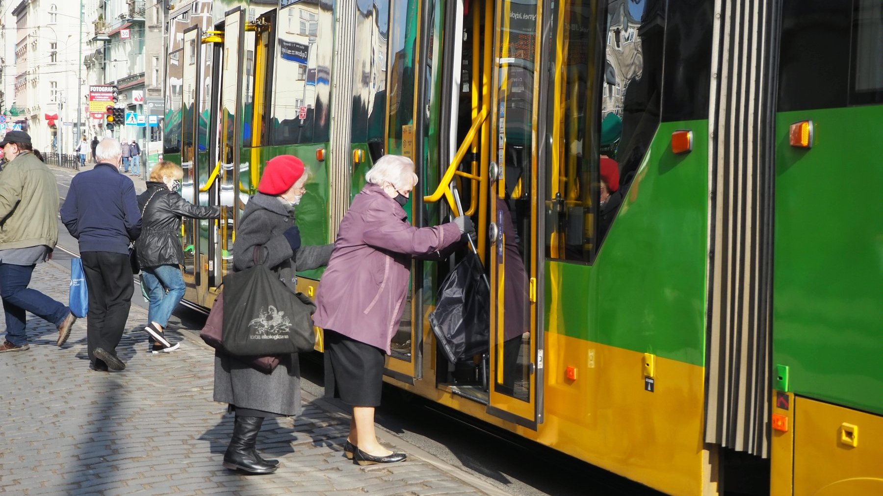 Zdjęcie przedstawia starsze osoby, które z trudem wsiadają do tramwaju na przystanku Rynek Jeżycki