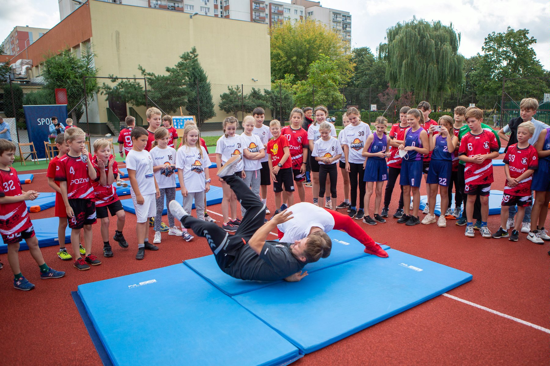 Na zdjęciu boisko szkolne, na macie dwóch sportowców, wokół dzieci - grafika rozmowy