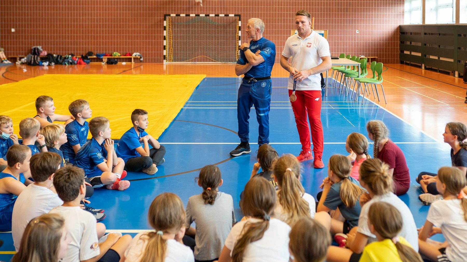 Sala gimnastyczna, na środku medalista olimpijski, wokół siedzą dzieci